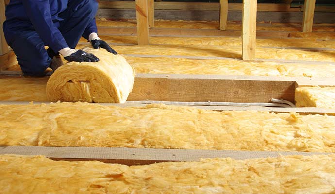 Worker installing yellow fiberglass insulation between wooden joists